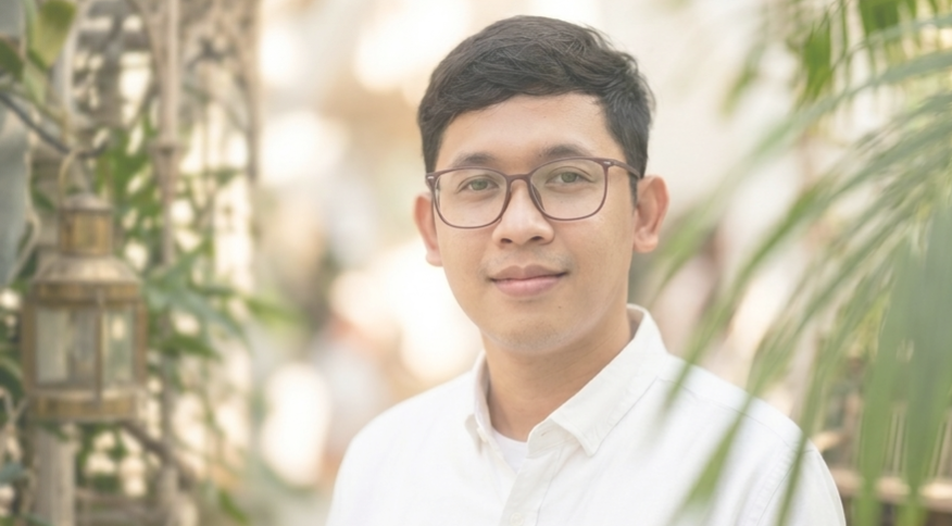 Potret Yossi mengenakan kemeja putih dan kacamata dengan senyum ramah, dikelilingi tanaman hijau yang artistik.